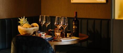 A dimly lit restaurant with a round table set for two, a bowl of fruit, wine glasses, and a bottle of wine.