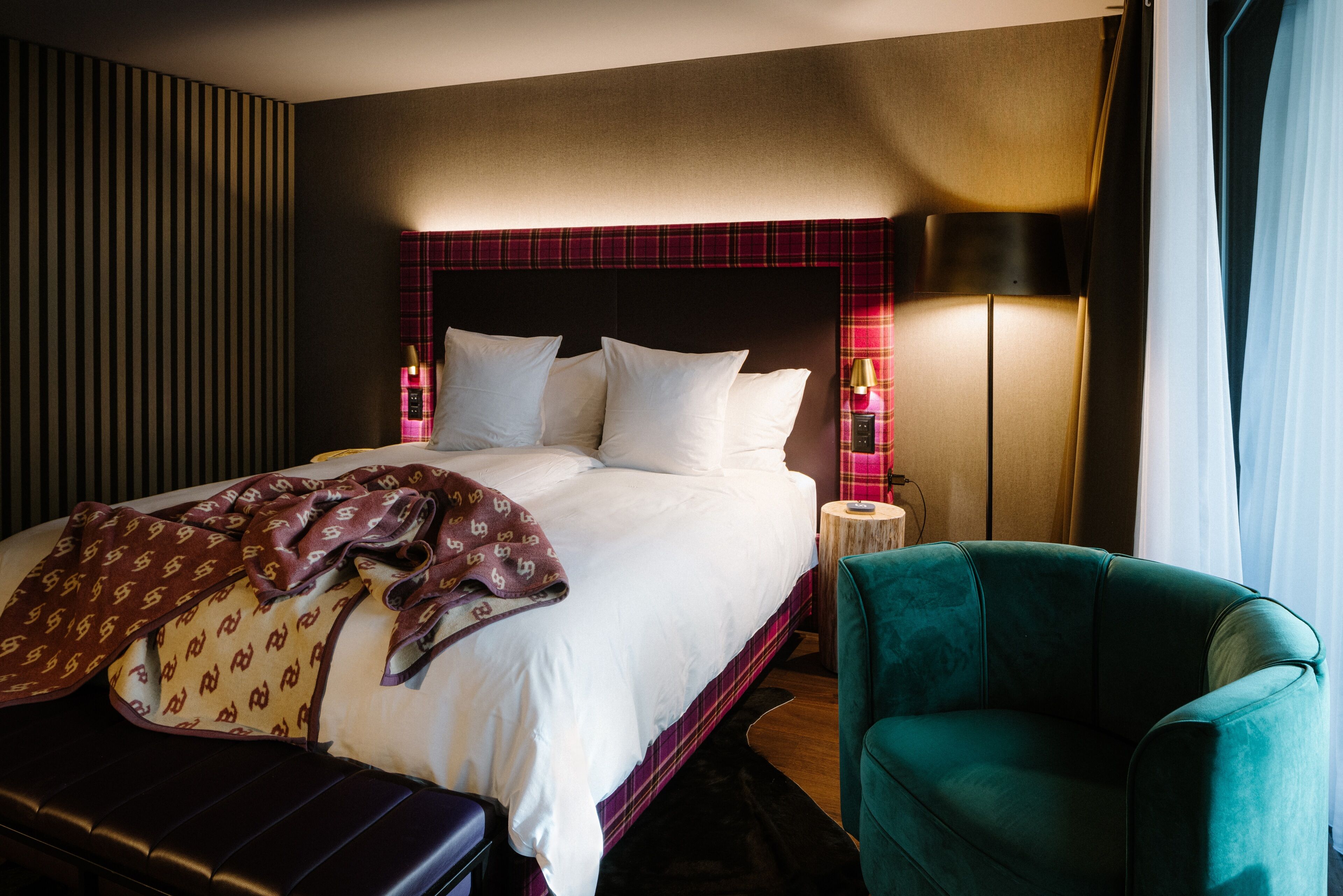 A hotel room with a bed, a green armchair, a nightstand with a lamp, and a striped wall.