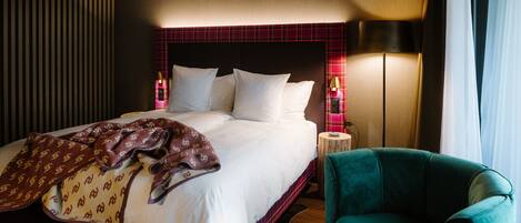A hotel room with a bed, a green armchair, a nightstand with a lamp, and a striped wall.