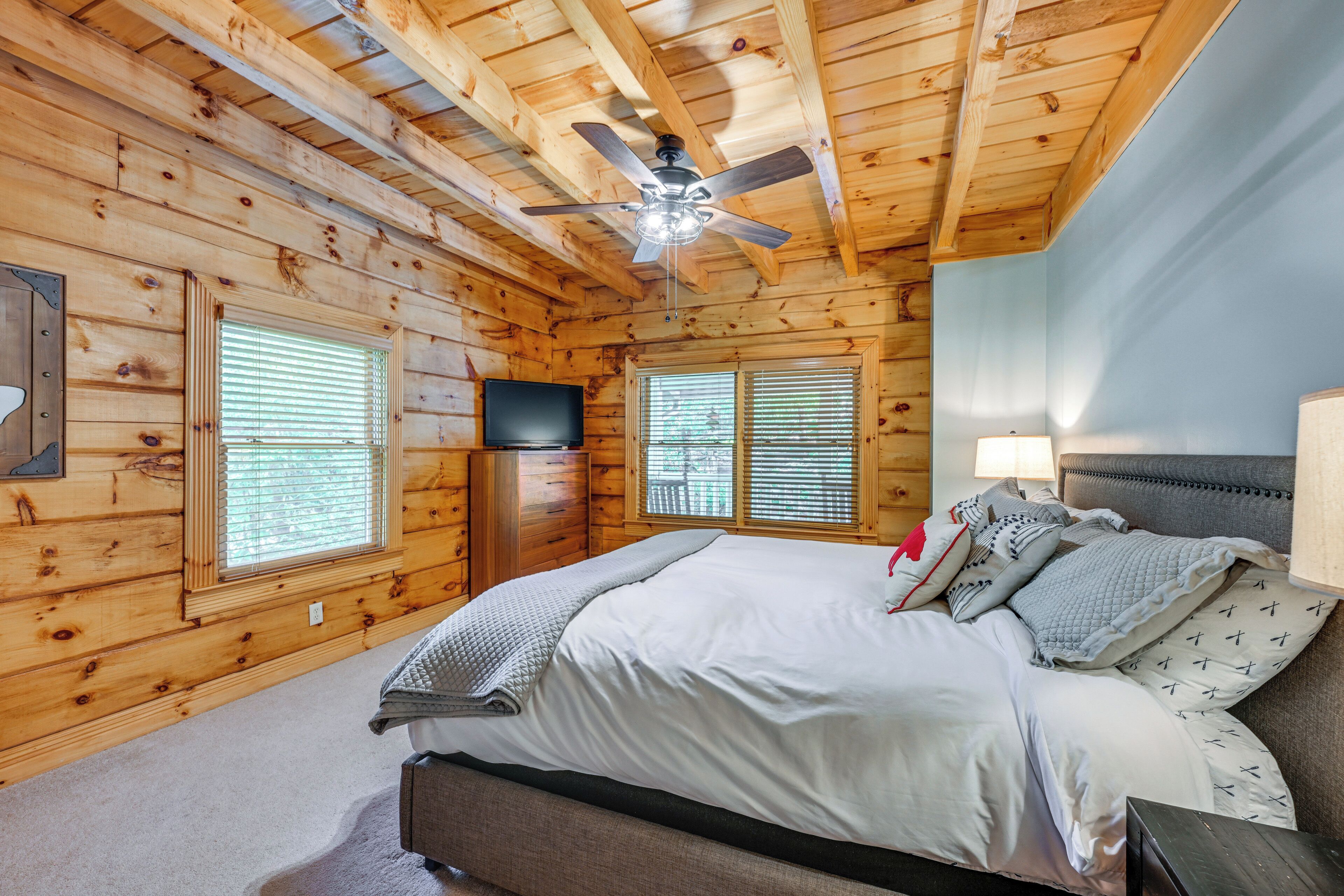 A log cabin bedroom with a bed, a ceiling fan, a TV, and two windows with blinds.