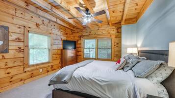 A log cabin bedroom with a bed, a ceiling fan, a TV, and two windows with blinds.