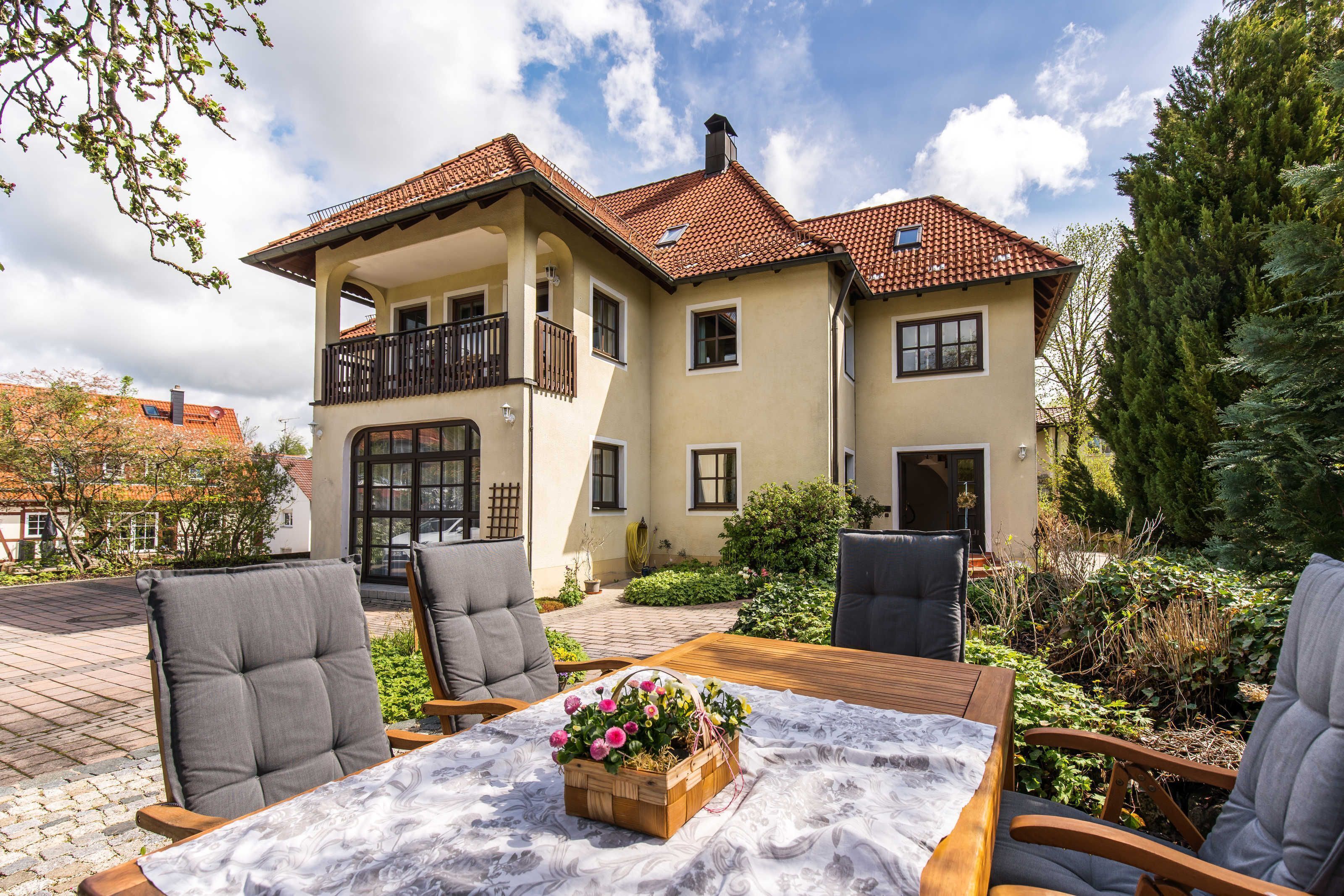 A two-story house with a balcony, a patio with outdoor furniture, and a garden.