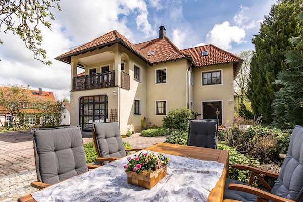 Una casa de dos pisos con un balcón, un patio con mobiliario exterior y un jardín.