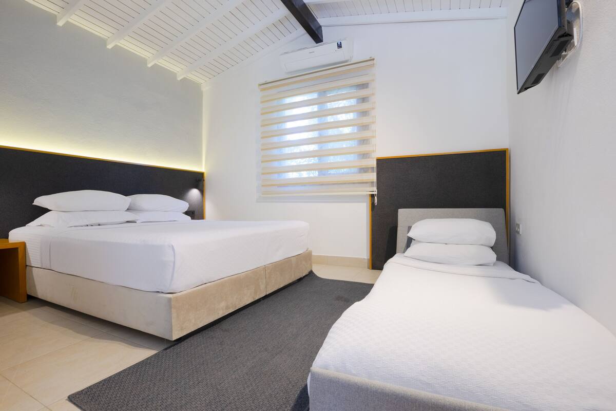 Una habitación de hotel moderna con dos camas, un ventanal con persianas, un televisor de pantalla plana montado en la pared y un techo con vigas a la vista.