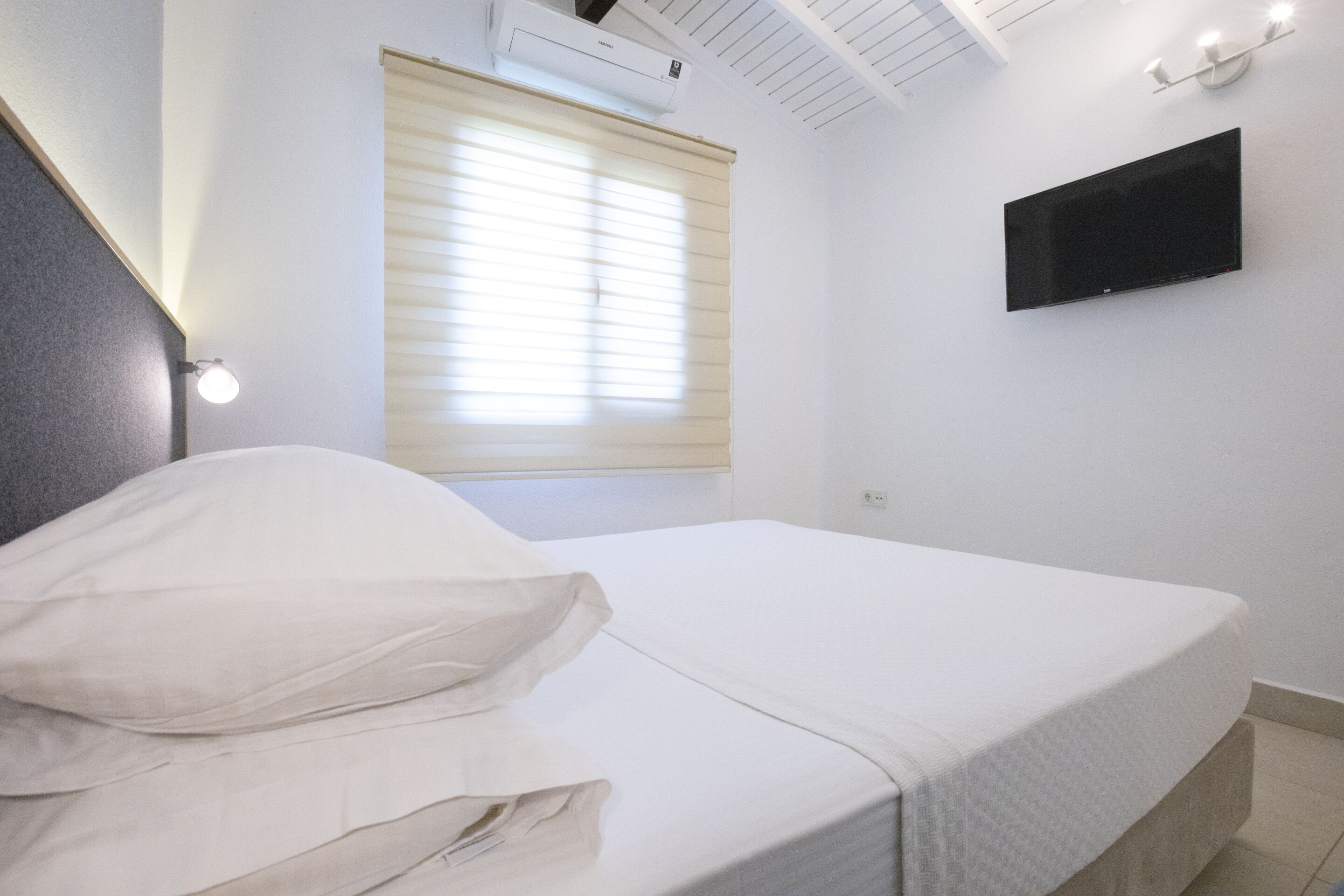 A bedroom with a bed, a window with blinds, a wall-mounted TV, and a ceiling fan.