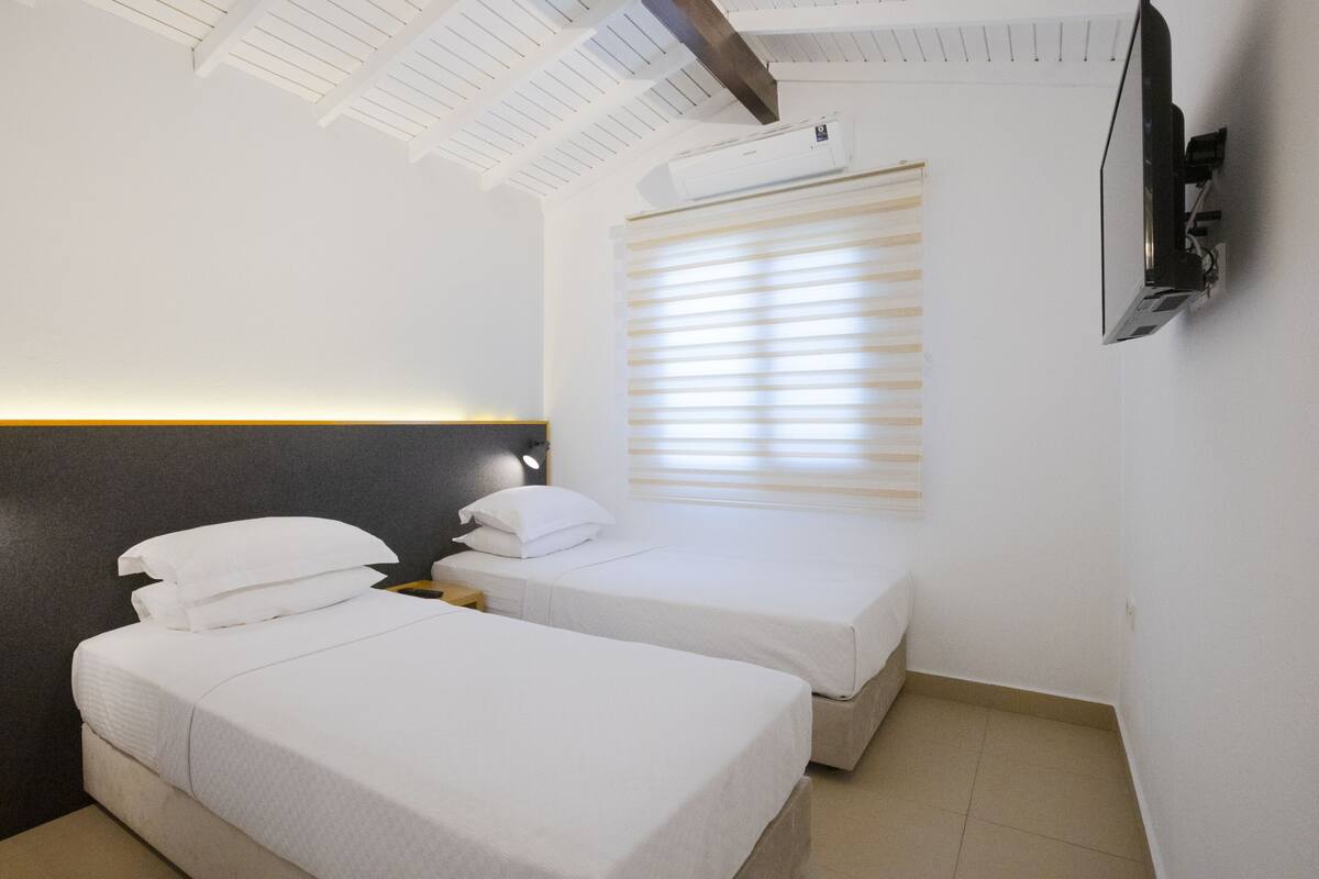 Habitación de hotel con dos camas, cabecera, ventana con persianas, unidad de aire acondicionado y televisión montada.