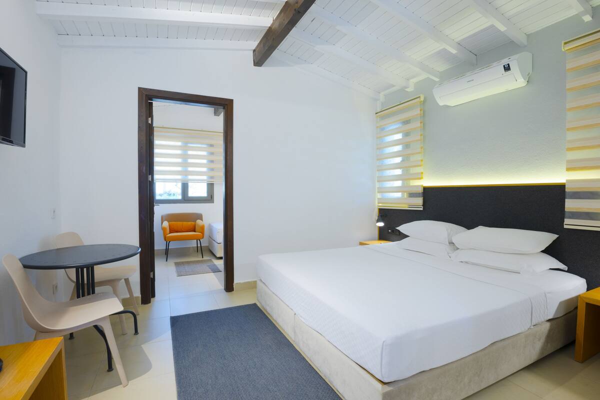 Una habitación de hotel moderna con una cama grande, una mesita pequeña y una silla. Se ve una puerta que conduce a otra habitación y un equipo de aire acondicionado en la pared.