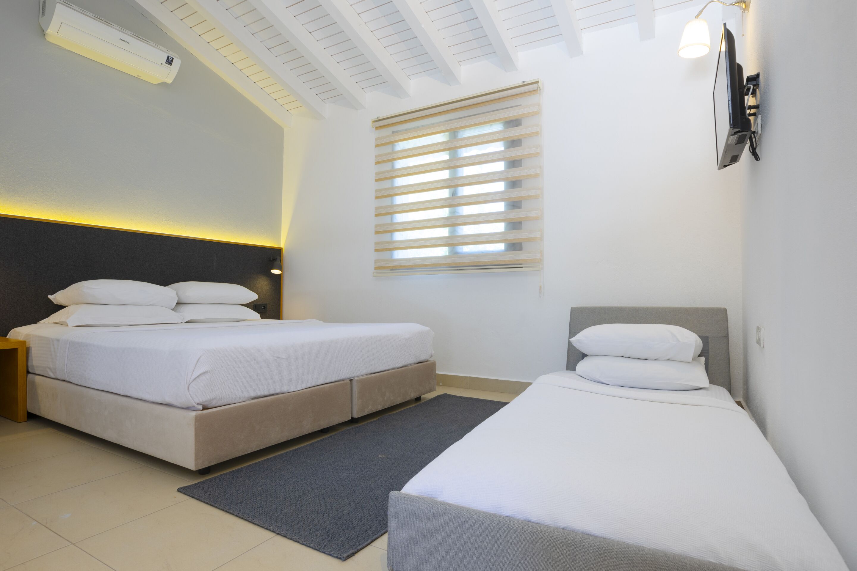 A modern hotel room with two beds, a ceiling fan, a wall-mounted TV, and a window with blinds.