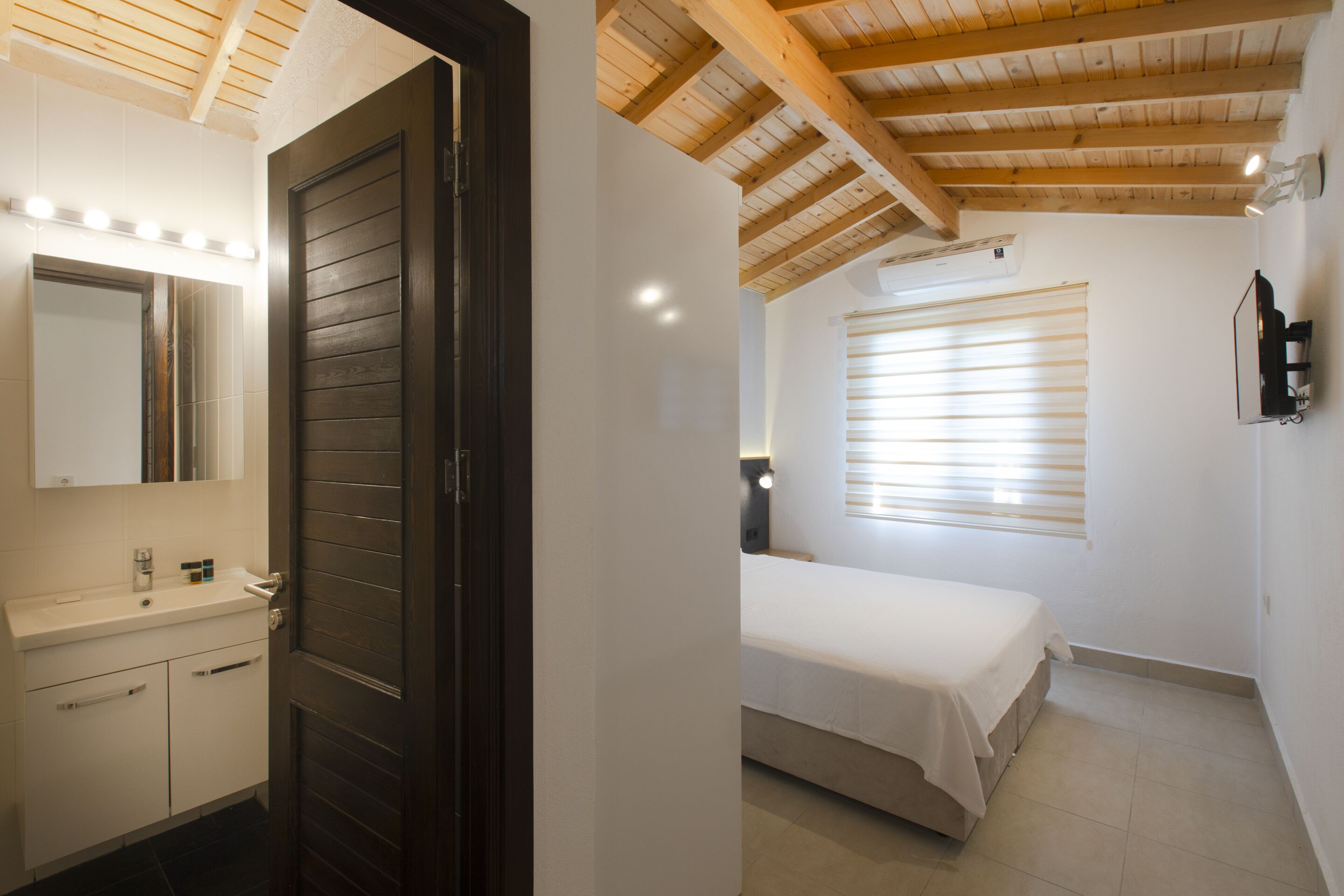 A bedroom with a bed, a bedside table, a window with blinds, and a wall-mounted TV.