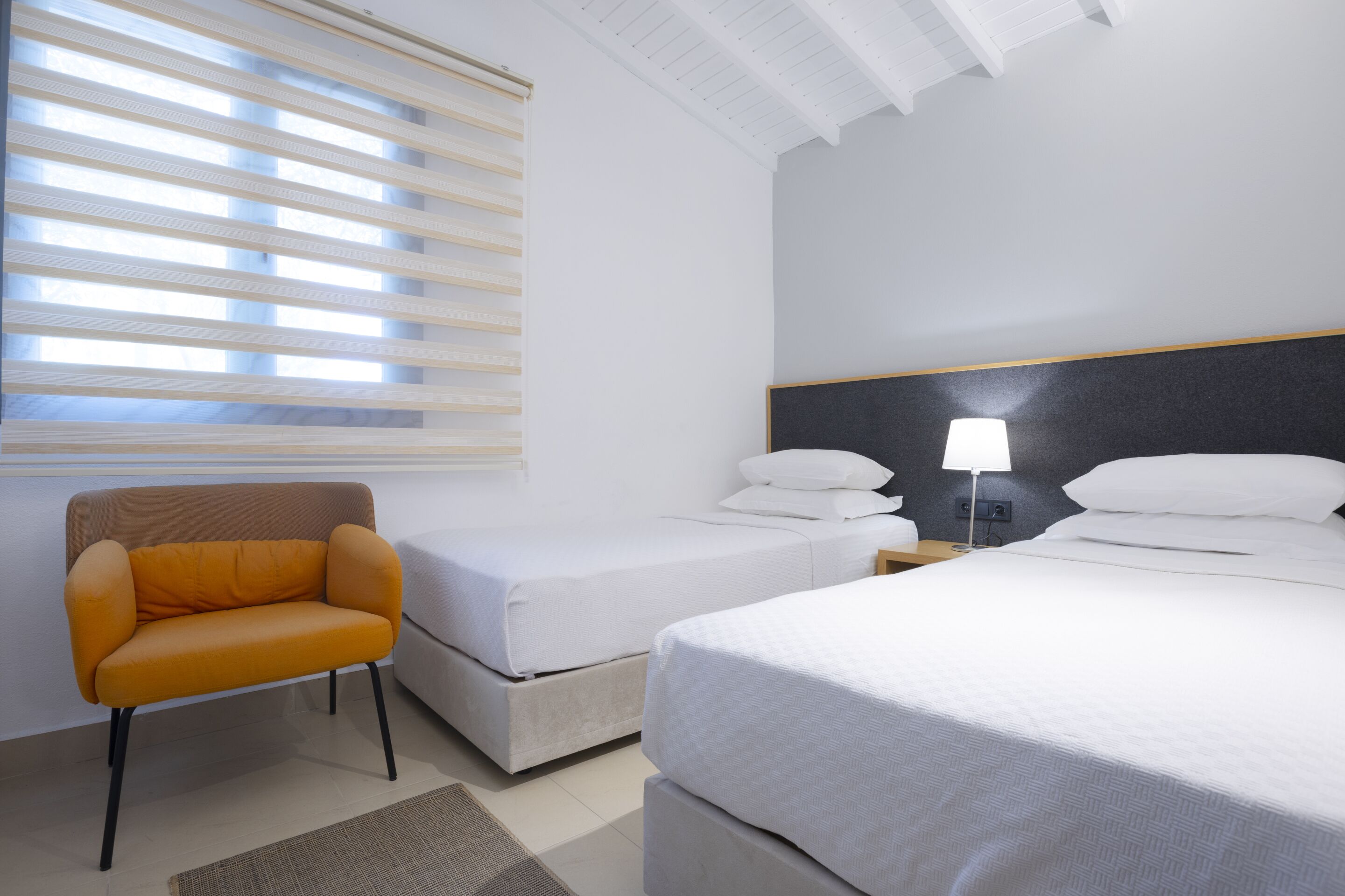 A hotel room with a single bed, a smaller bed, a chair, a nightstand with a lamp, and a window with blinds.