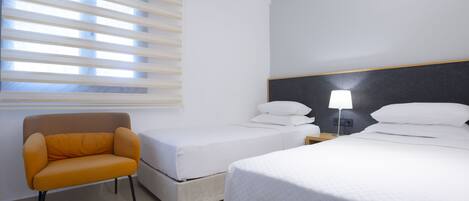 A hotel room with a single bed, a smaller bed, a chair, a nightstand with a lamp, and a window with blinds.