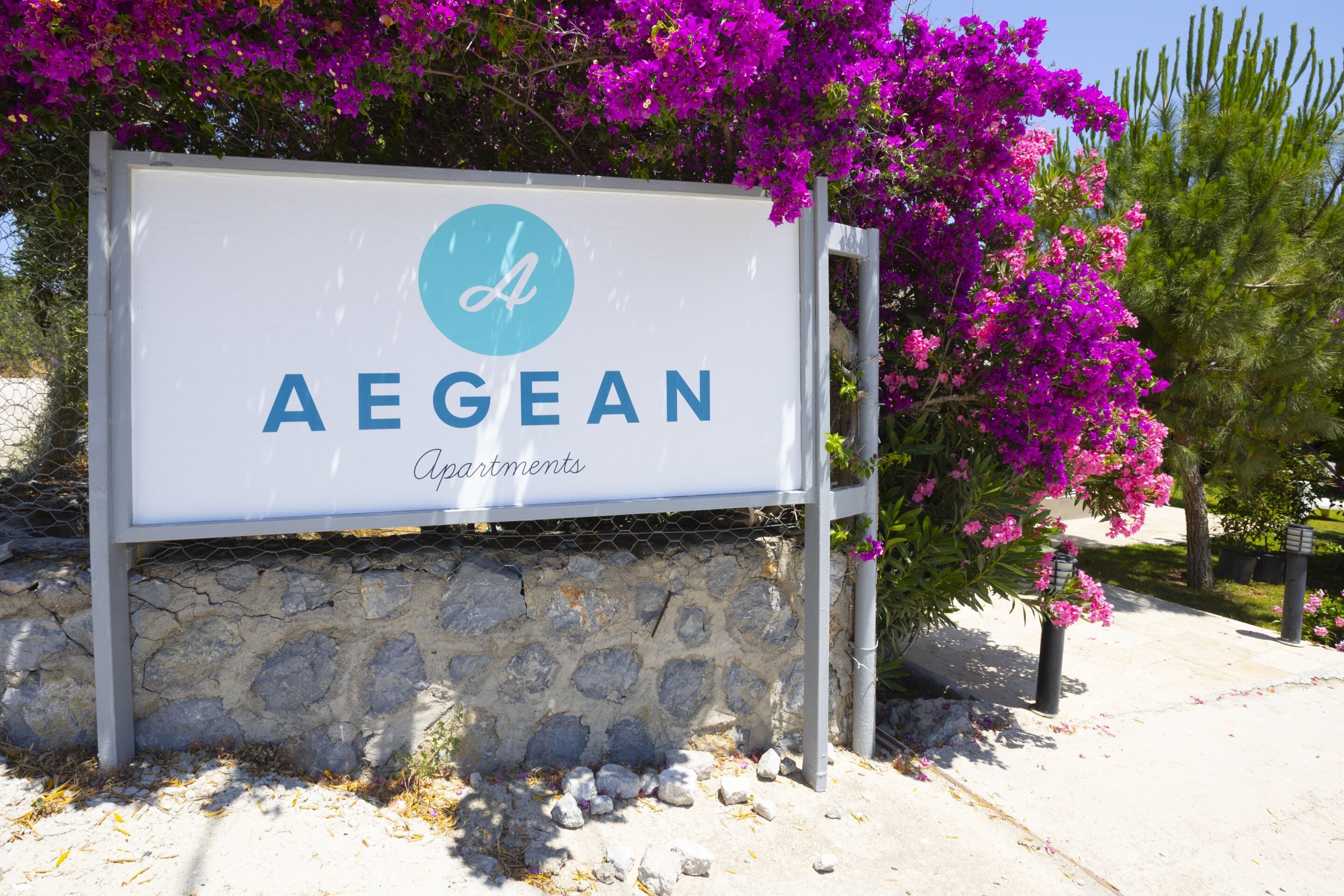 Een bordje voor Aegean Apartments, met een logo, voor een achtergrond van paarse bougainvillea en een stenen muur.
