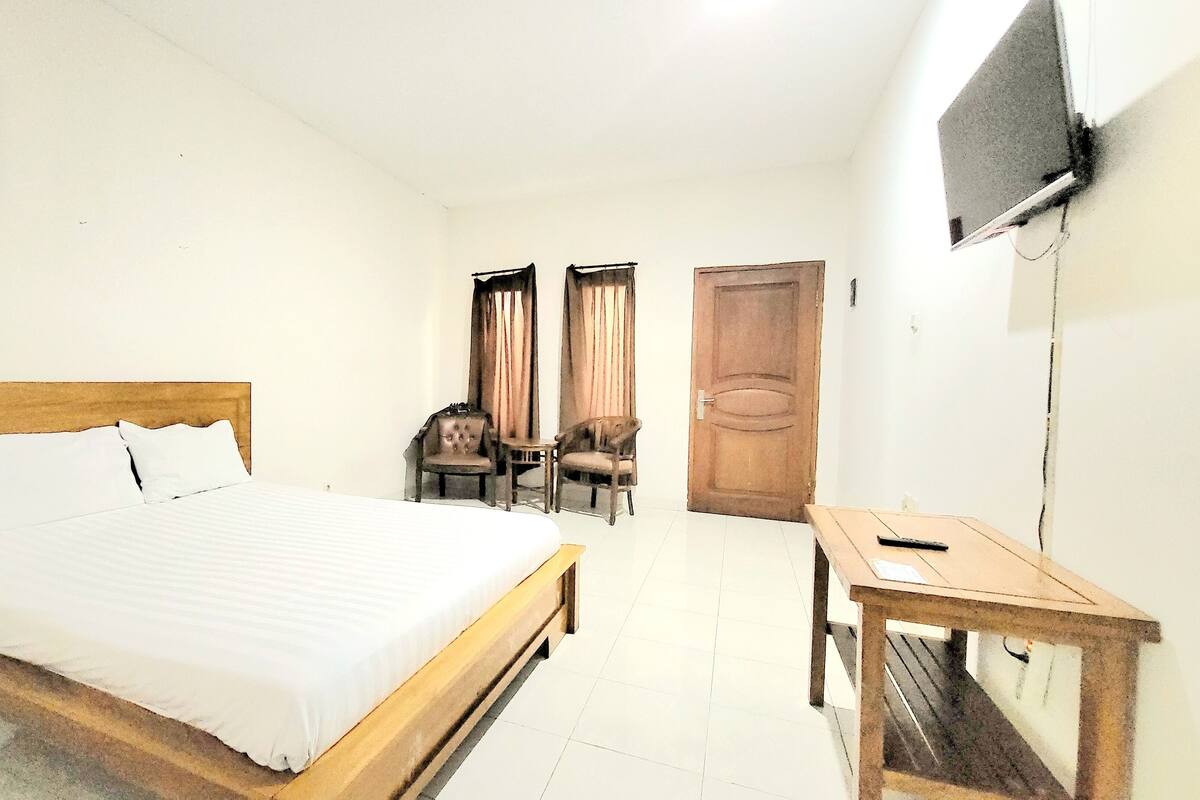 A hotel room with a wooden bed, a small wooden table, two chairs, a TV mounted on the wall, and a door.