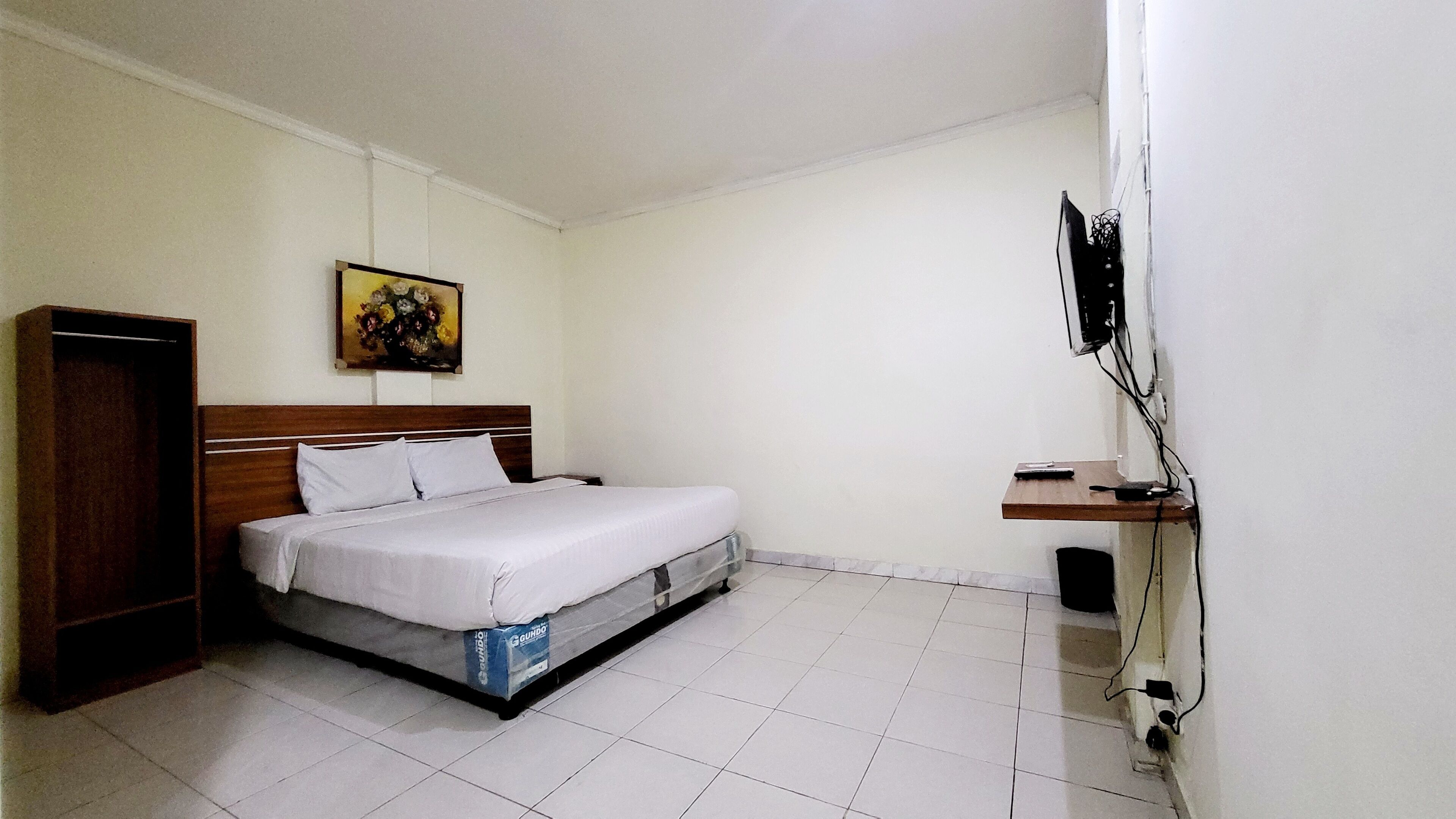 A bedroom with a bed, a wooden wardrobe, a small table, a TV mounted on the wall, and a painting above the bed.