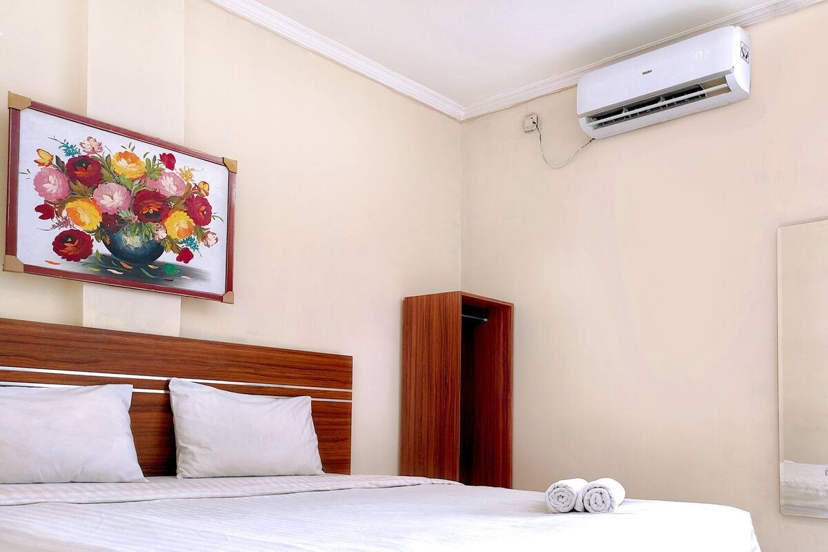 A hotel room with a bed, a painting, an air conditioning unit, a wardrobe, and a mirror.