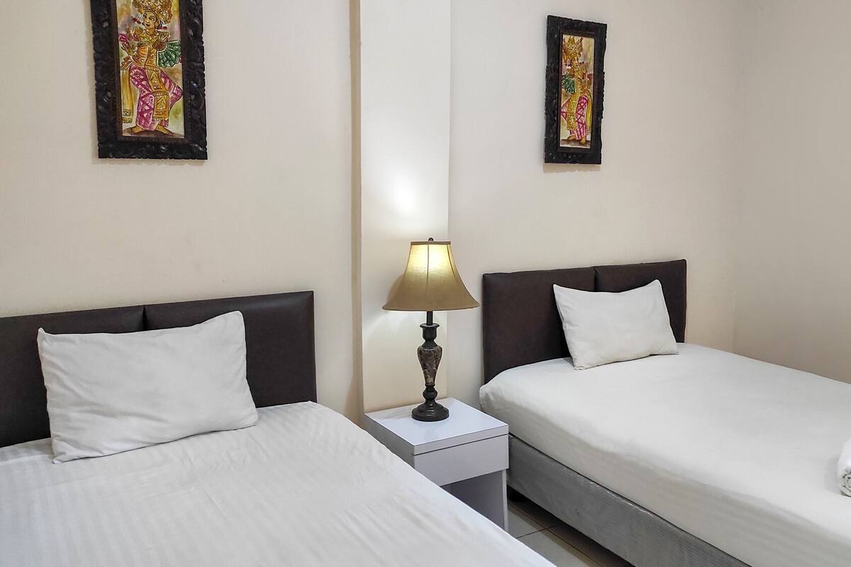 A hotel room with two single beds, a nightstand with a lamp, and two wall-mounted paintings.