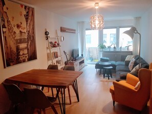 A living room with a wooden dining table, a sofa, and a TV. There is a bookshelf with books and a guitar. A large painting is on the wall, and a window provides a view of the outside.