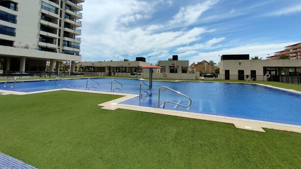 A swimming pool with a surrounding grassy area, a building with multiple balconies, and a clear sky. - NON STRESS First Level (Alboraya)