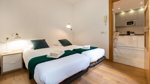 A hotel room with a bed, bedside table, lamp, and a view of the kitchen area. - Fantastic and Bright Apartment in the Heart of Gothic area 10 min from the beach (Barcelona)