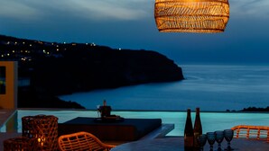 An outdoor dining area with wicker furniture, a table with bottles and glasses, and a view of the sea at dusk. - Ambeli Sunset Villas (Santorini)