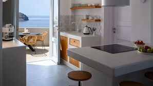 A modern kitchen with a large island, wooden stools, and a view of the sea through an open door. - Ambeli Sunset Villas (Santorini)