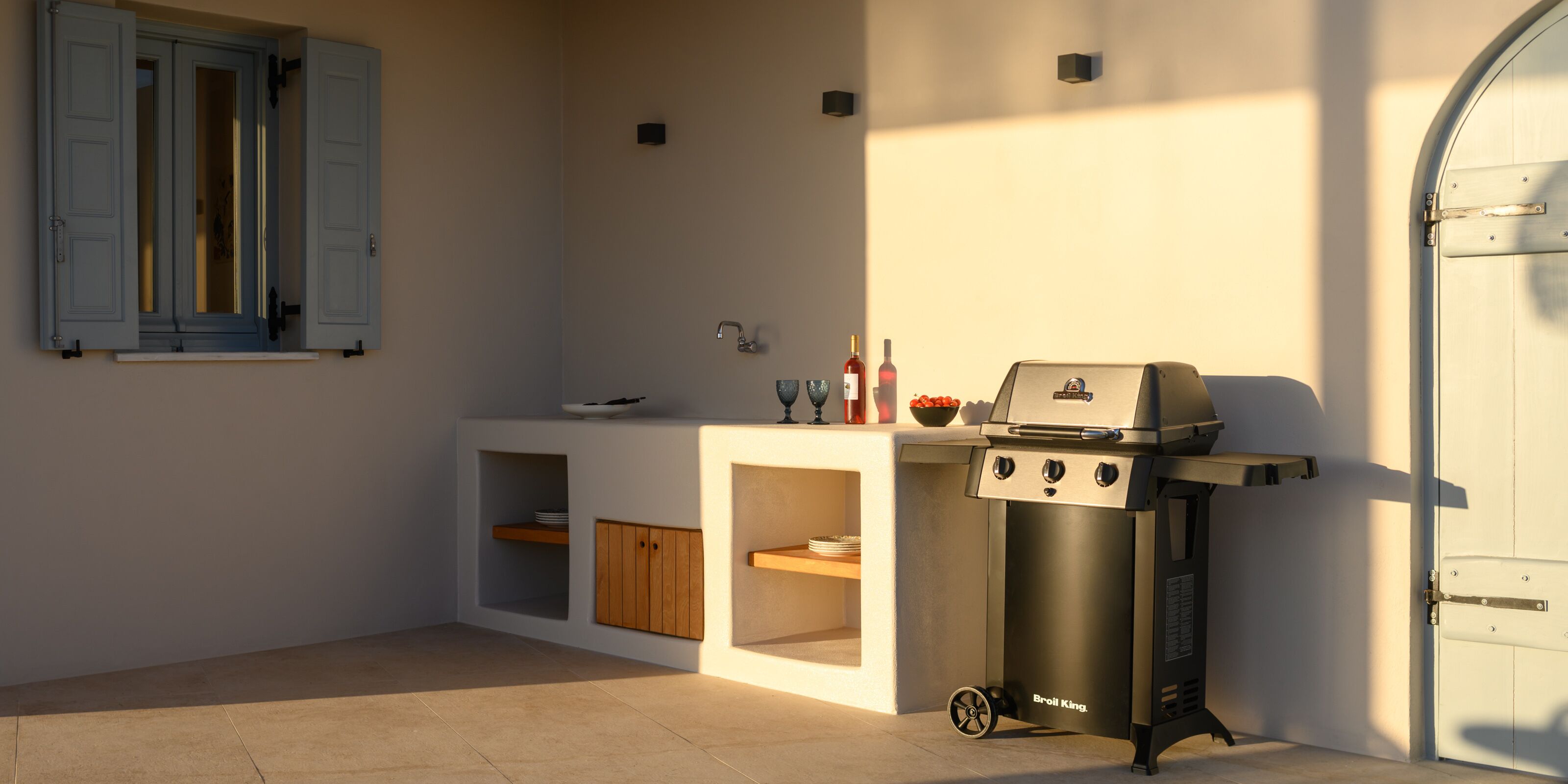 A compact kitchen with a built-in grill, a window with shutters, and a small countertop with a bowl and bottles.