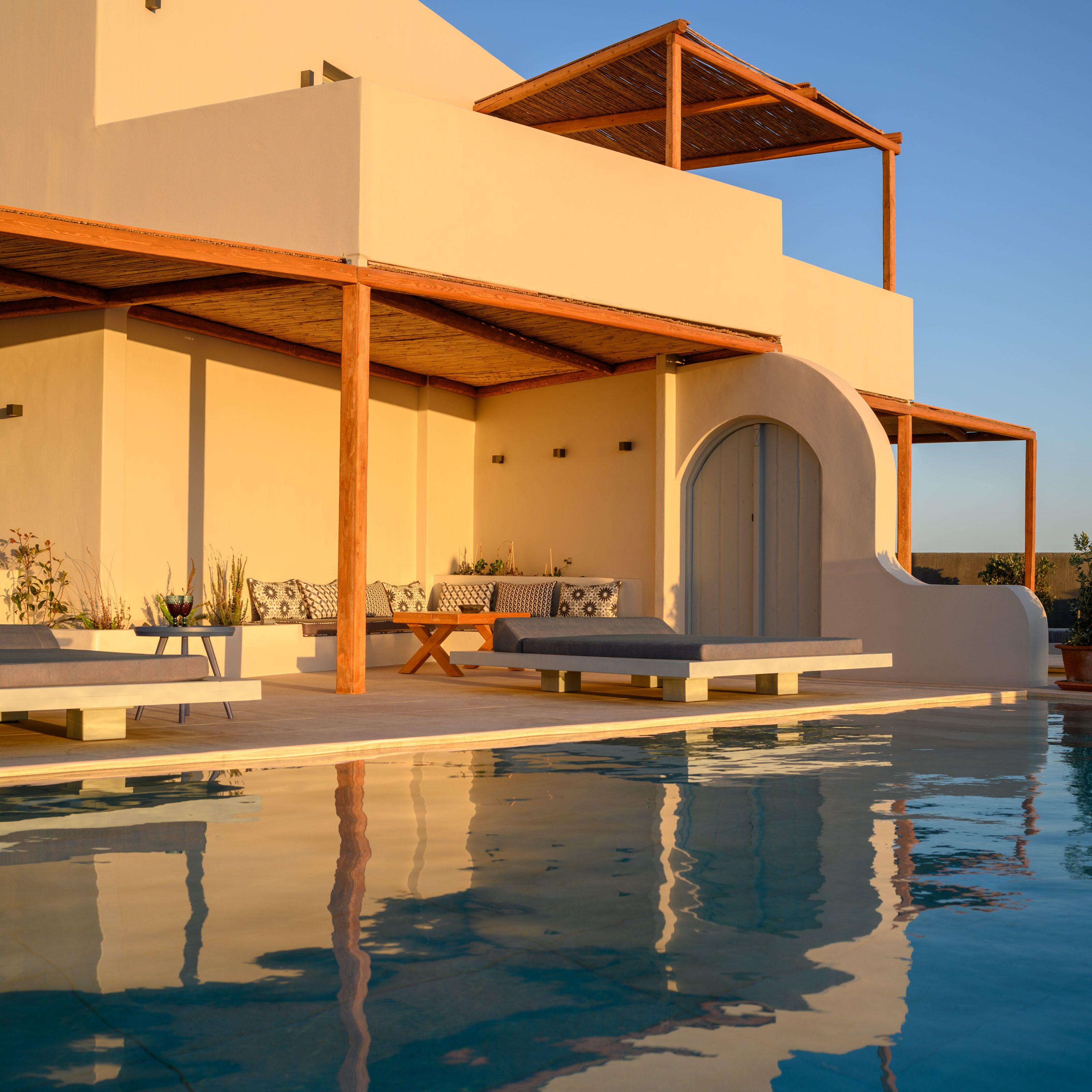 Un área moderna junto a la piscina con una pérgola de madera, una zona de estar con cojines y una piscina cristalina que refleja el entorno.