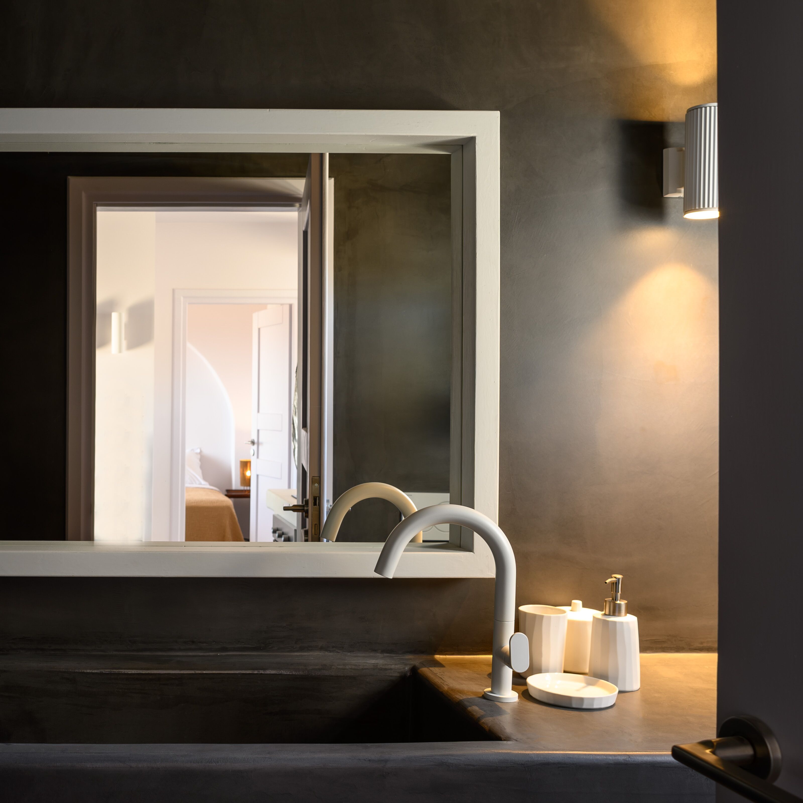 Ein Badezimmer mit einem dunklen Holzwaschtisch, einem weißen Waschbecken und einem großen Spiegel. Eine Wandleuchte ist über dem Spiegel angebracht.