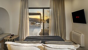 A modern hotel room with a large bed, a TV mounted on the wall, and a view of a snowy landscape through a large window. - Ambeli Sunset Villas (Santorini)