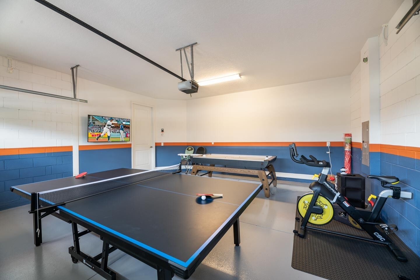 A ping pong table in a room with blue and orange walls, a treadmill, and a television displaying a sports event.