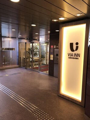 Une entrée d’hôtel moderne avec une porte en verre, un comptoir d’accueil et une enseigne indiquant le nom « VIA INN Hiroshima ».
