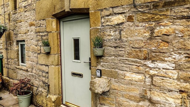 Un bâtiment en pierre avec une porte de couleur claire, de petites fenêtres et des plantes en pot.