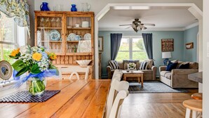 A cozy living room with a wooden cabinet filled with dishes, a dining table with chairs, a sofa, and a ceiling fan. - Robeson Retreat (Sag Harbor)