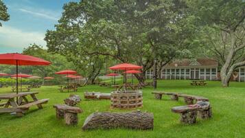 An outdoor area with picnic tables and red umbrellas, surrounded by trees.