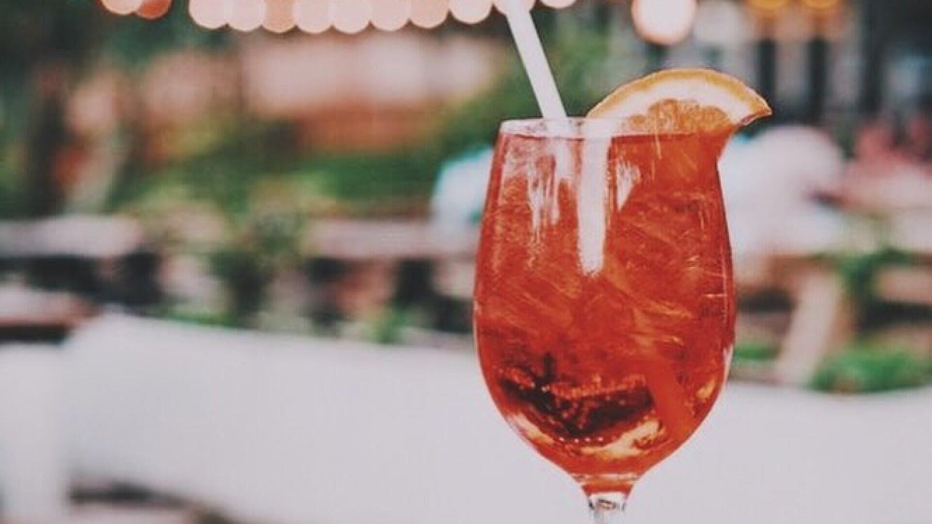 A glass of red beverage with a lemon slice and a straw, set against a blurred background of a restaurant or bar setting.