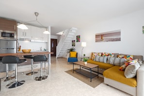 A modern kitchen with a microwave, stove, and bar stools. There is a staircase leading to another level. A sofa with a mustard throw blanket and colorful pillows is in the living area.