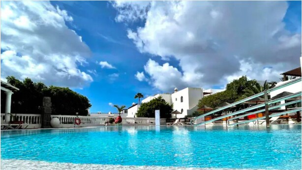 Una piscina azul cristalina rodeada de edificios blancos, palmeras y un cielo despejado.