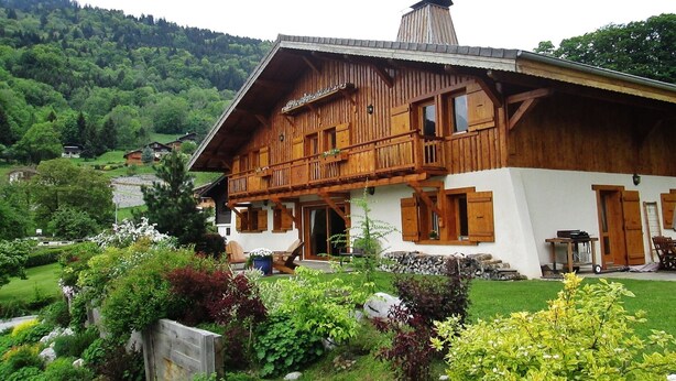 Een klassiek houten chalet met balkon, omgeven door weelderige begroeiing en een bergachtig landschap.