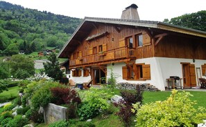 En traditionel træchalet med balkon, omgivet af frodig vegetation og et bjerglandskab.