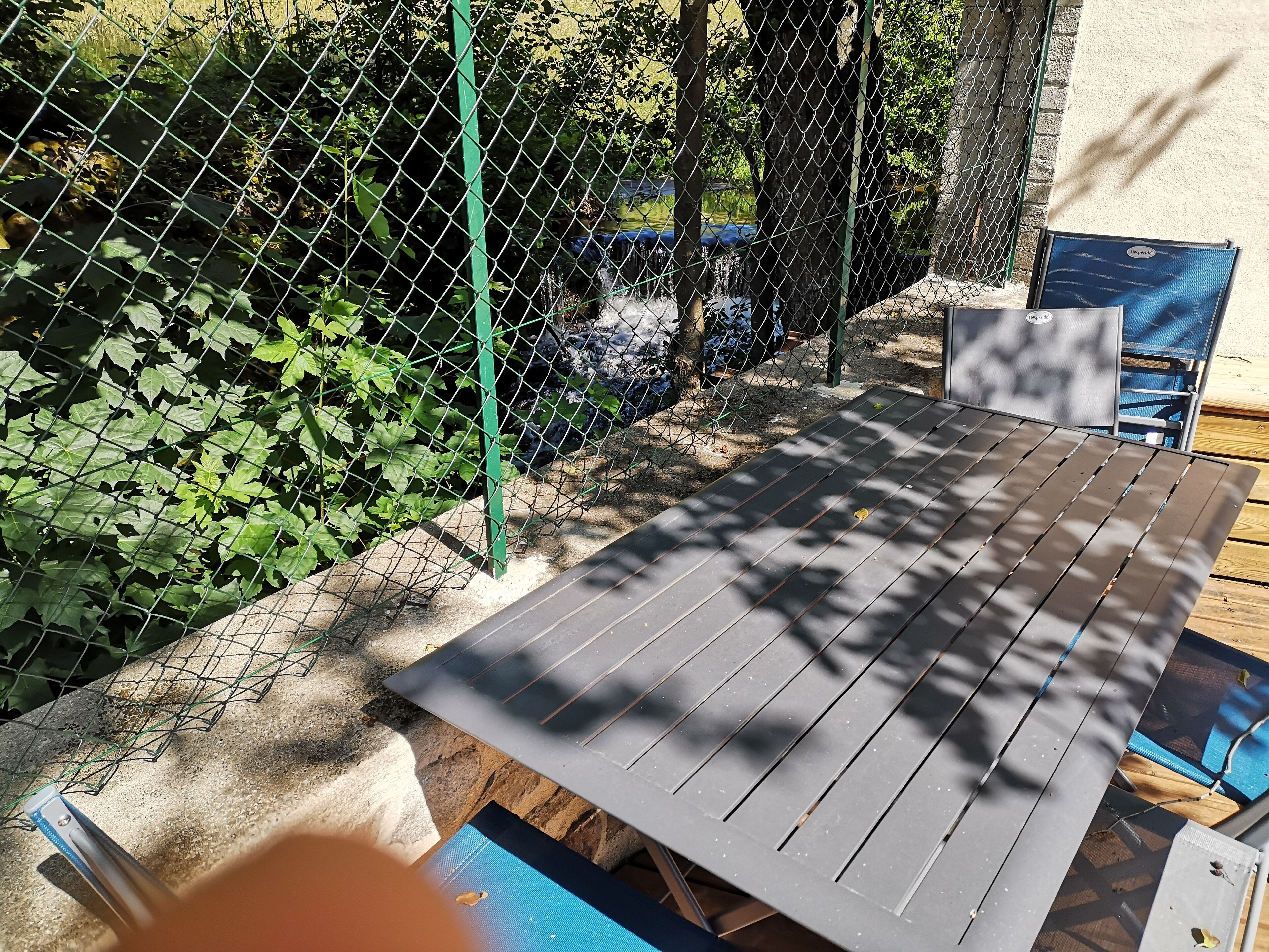 A patio area with a metal table and chairs, surrounded by a chain-link fence and greenery.