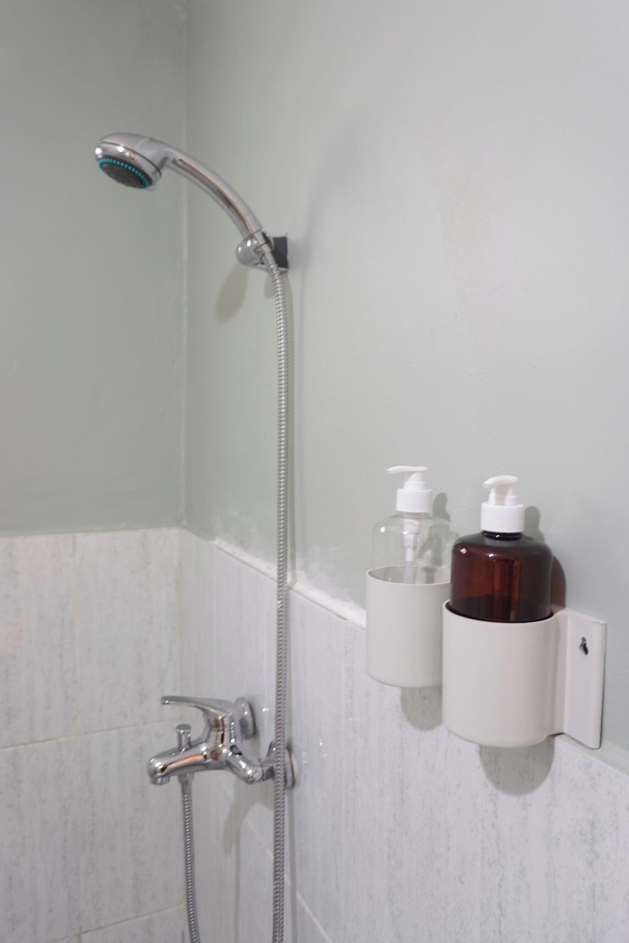 Une douche équipée d’un pommeau de douche à main et d’un robinet mural. Deux flacons de savon liquide sont fixés au mur.