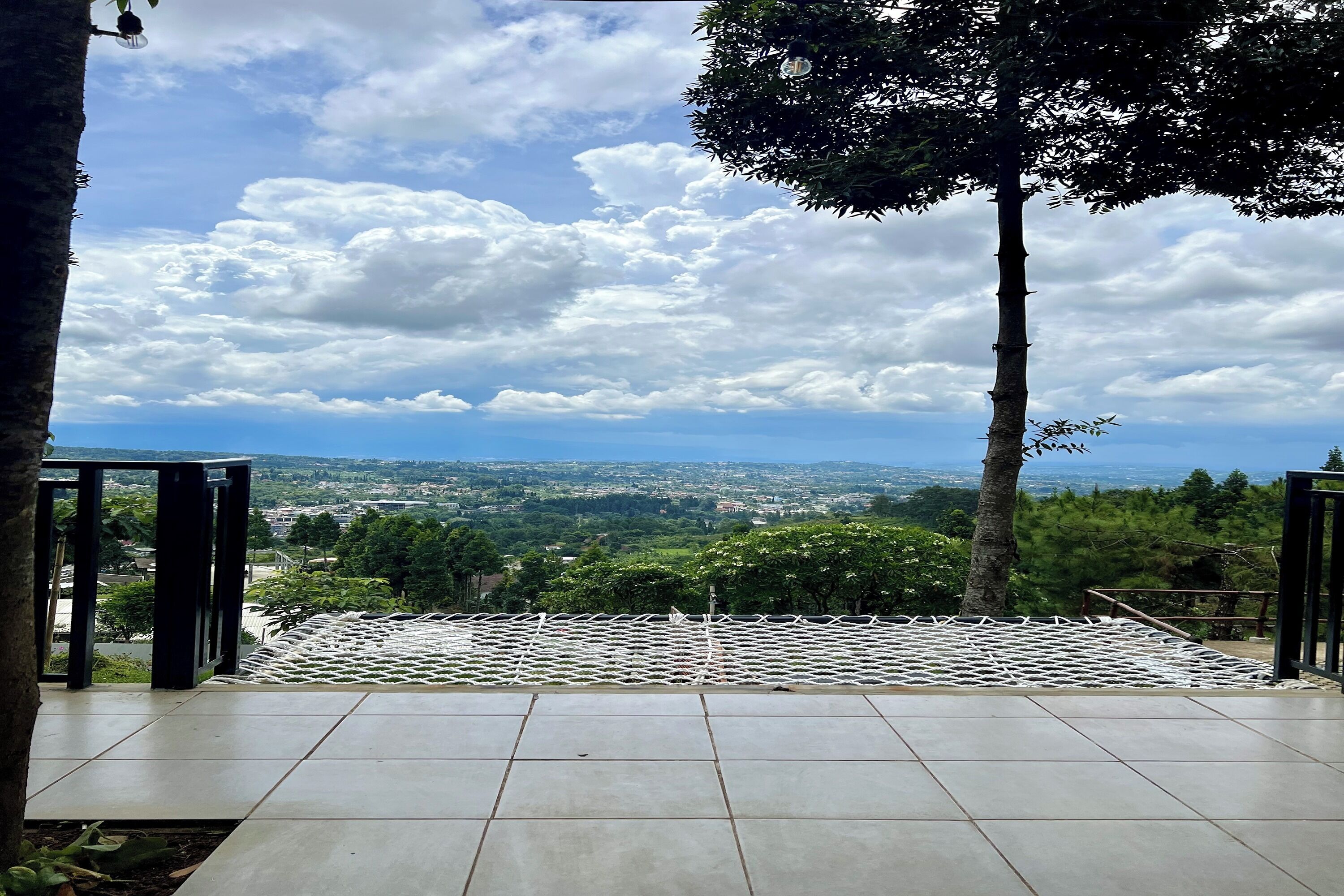 A terrace with a view of a cityscape, surrounded by trees and a white mesh barrier.
