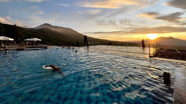 Uma piscina com água cristalina, rodeada por um deck com guarda-sóis e pessoas.