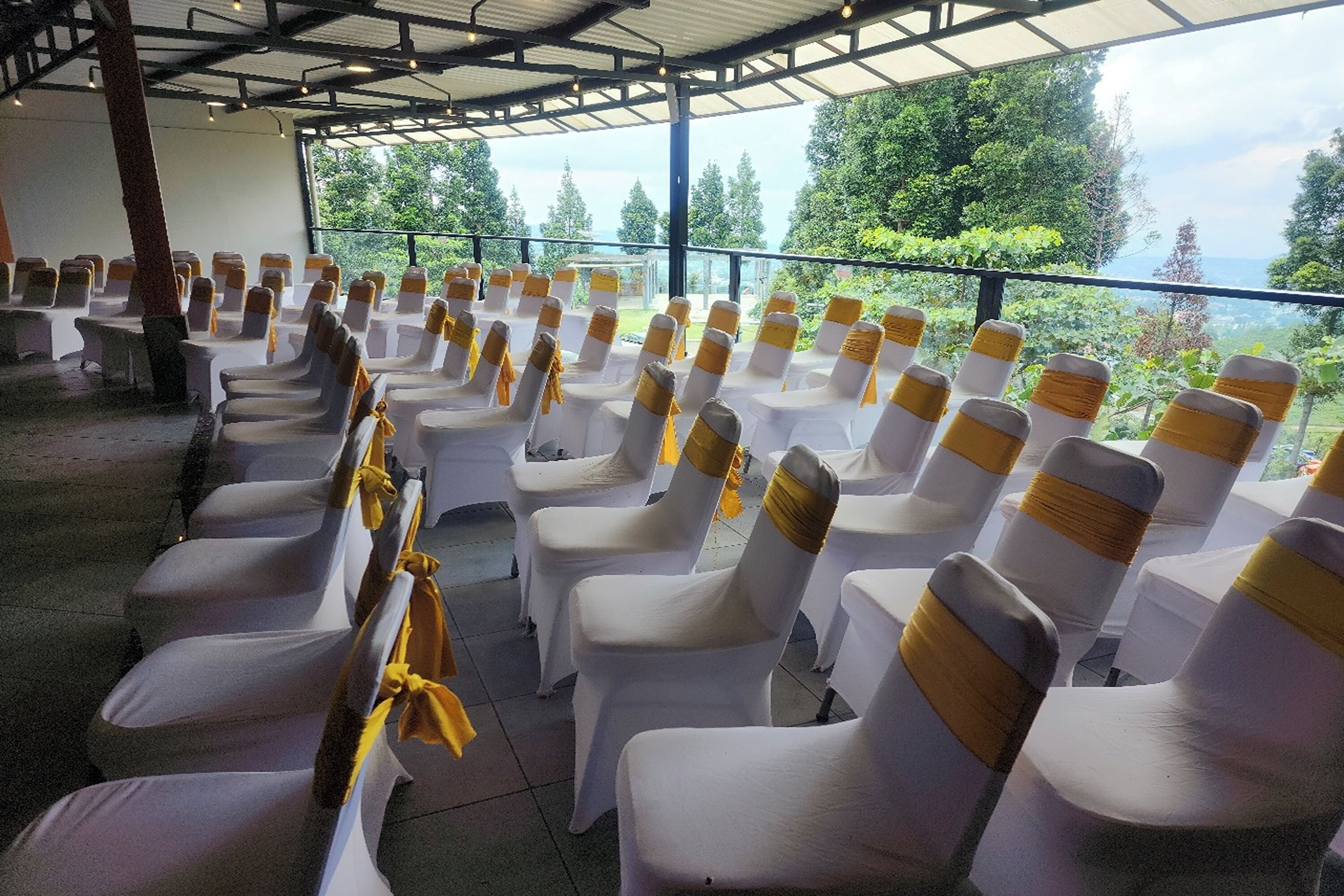 Rows of white chairs with yellow sashes, arranged in a covered outdoor area with a view of trees and a distant landscape.