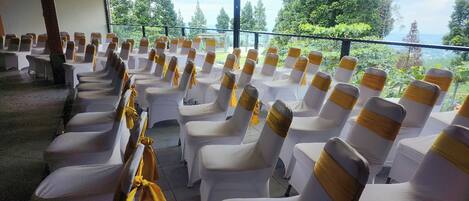 Rows of white chairs with yellow sashes, arranged in a covered outdoor area with a view of trees and a distant landscape.