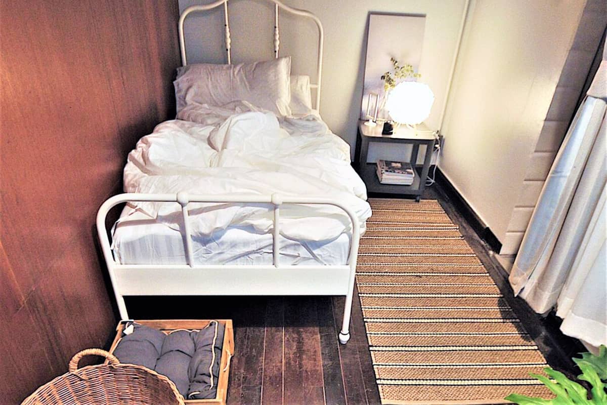 A neatly made bed with white linens, a bedside table with a lamp, a wicker basket, and a striped rug on a wooden floor.