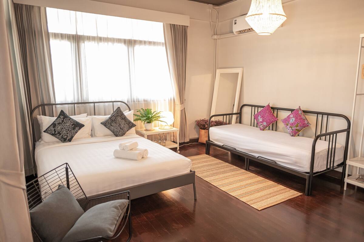 A room with two beds, a bedside table with a lamp, a mirror, and a window with curtains.