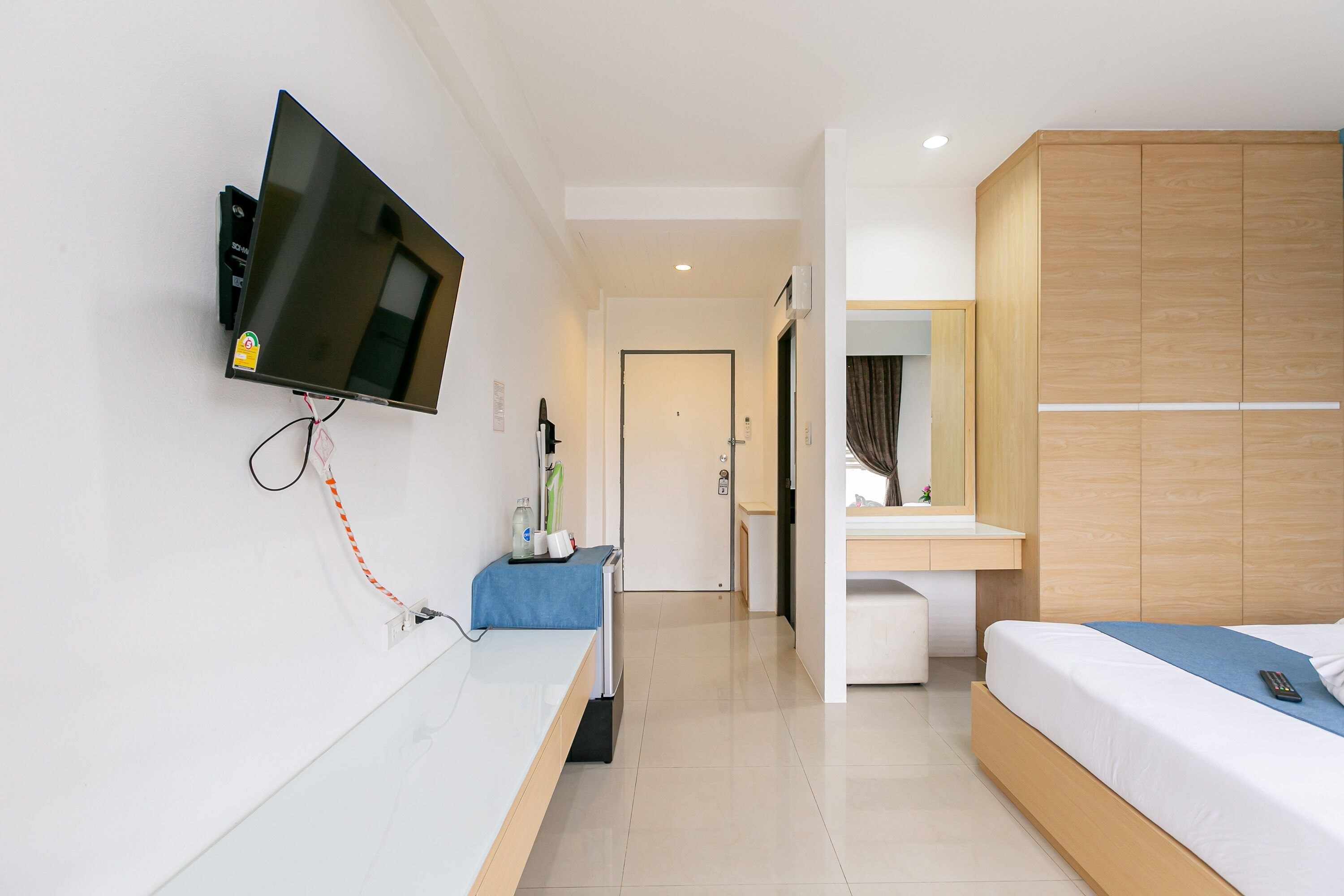 A modern hotel room with a flat-screen TV mounted on the wall, a bedside table with a remote control, a bed with a blue bedspread, a wooden wardrobe, and a bathroom visible in the background.