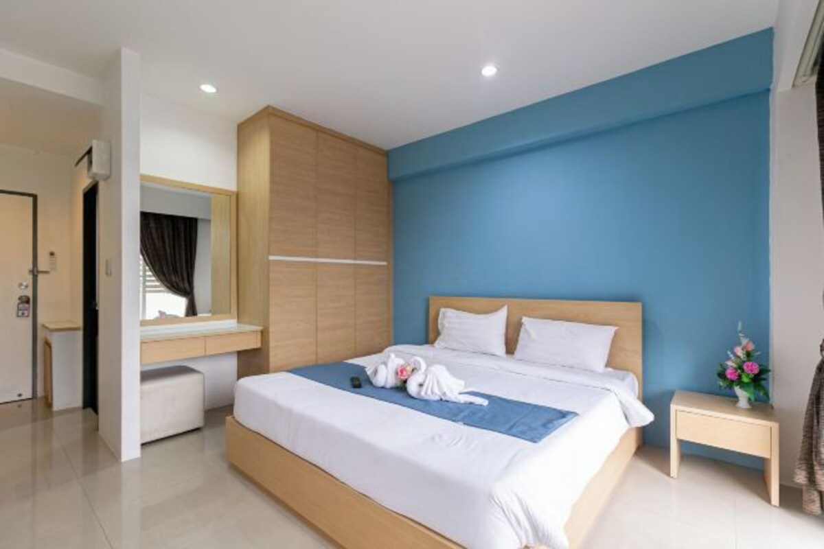 A modern hotel room with a large bed, a wooden headboard, a bedside table with a vase of flowers, and a mirror above the bed.