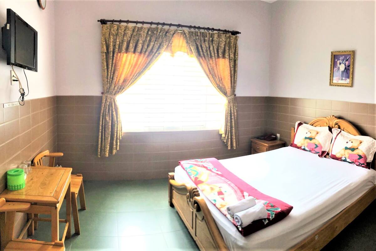 A bedroom with a bed, a wooden nightstand, a table with chairs, a TV mounted on the wall, and a window with curtains.
